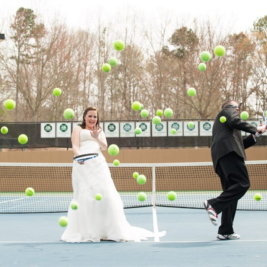 Bride + Groom on tennis court laughing as dozens of tennis balls come at them
