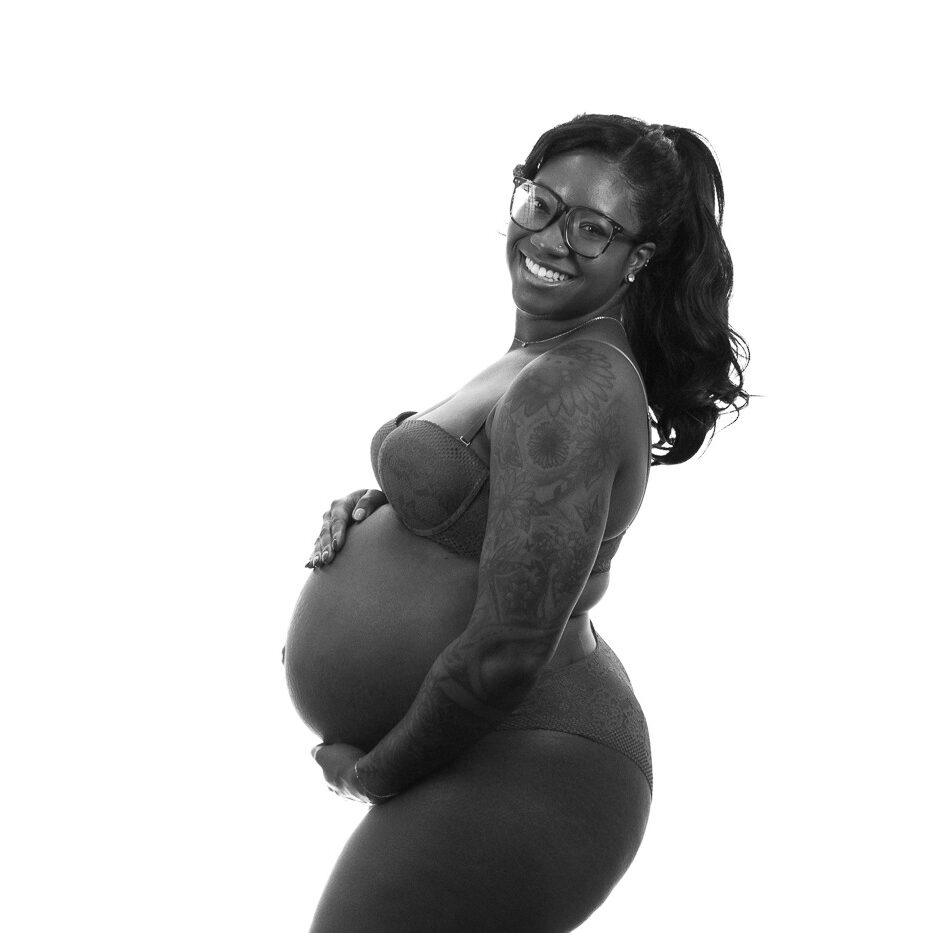 Black and white photograph of a pregnant momma with a long pony tail holding her belly and smiling