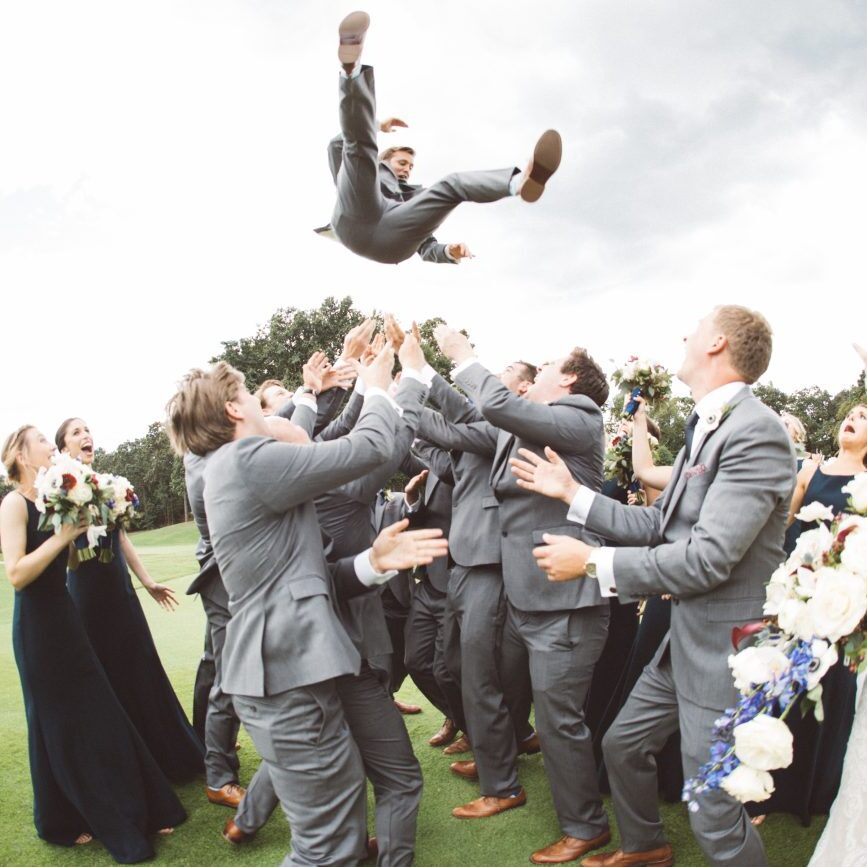 Documentary wedding photographer captures spontaneous moment of groom being thrown into the sky above groomsmen while bride and bridesmaids watch in surprise outside of the Atlanta Country Club for a wedding reception