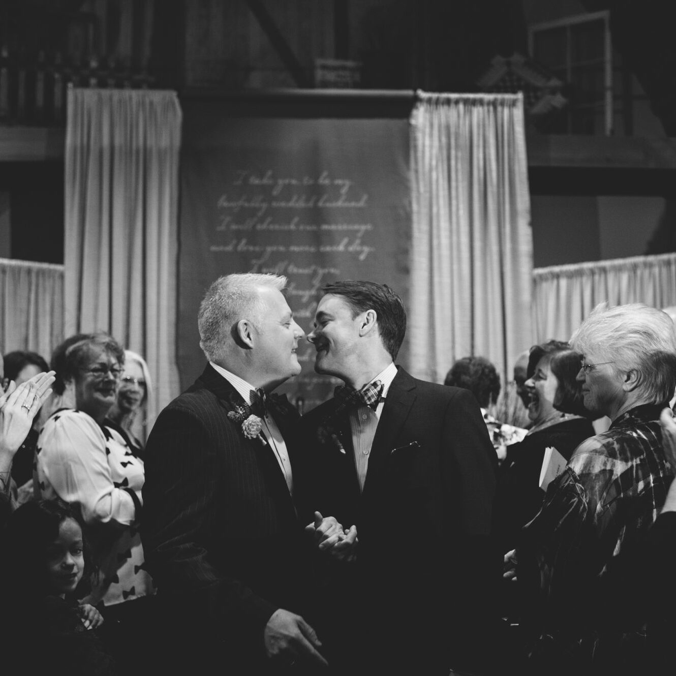 Black and white photograph of LGBT wedding with two grooms about to kiss after their ceremony and stopping in the aisle