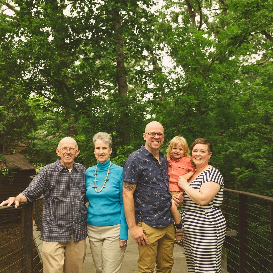 Multi-generational photo with grandparents standing together on a bridge with tall green trees behind them