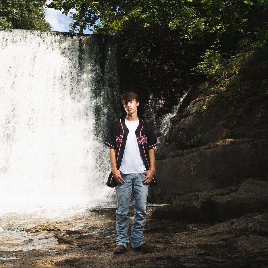 High school senior boy standing with thumbs in jeans pockets. Roswell Mill dam waterfall behind him and wearing his Bulldogs baseball jersey