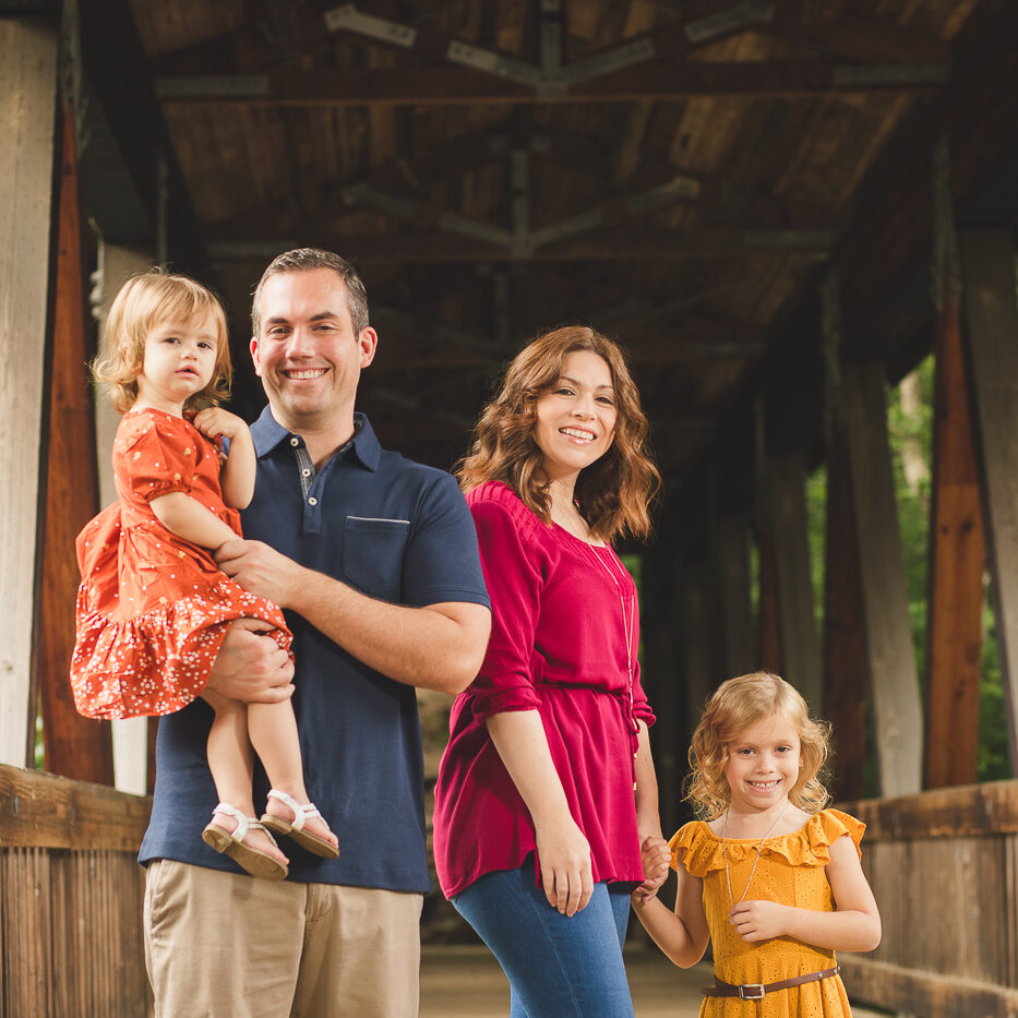 Gorgeous couple with two cute little girls under the age of five all dressed in bright colors and smiling for family photographs inside Roswell Mill covered bridge