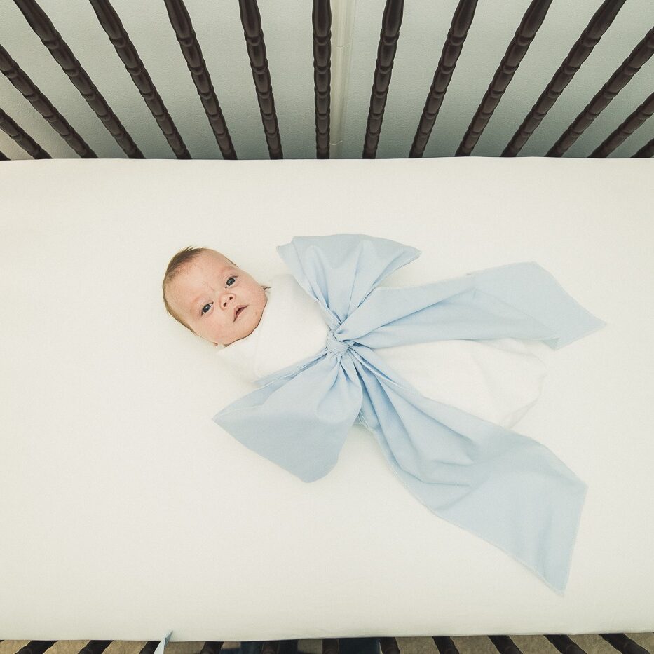 Newborn baby boy wrapped in a white blanket and big blue bow. Laying on his back looking up from the middle of his dark wood crib and white mattress