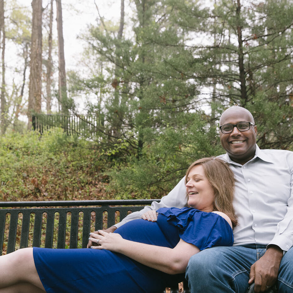 Beautiful pregnant mom in blue dress laying down on her husband's lap on a park bench