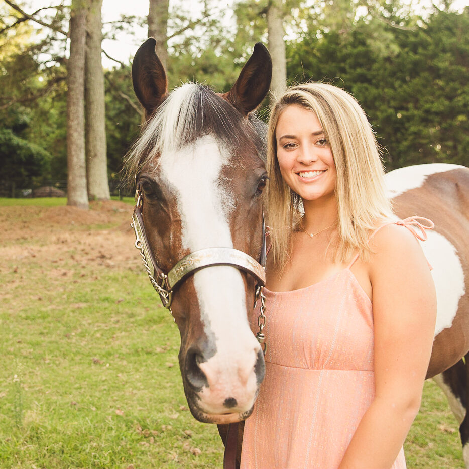 A blonde high school senior girl standing next to her horse and smiling