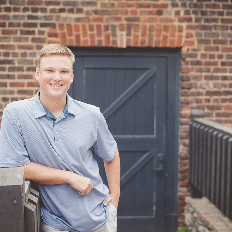 Smiling teenage boy in blue shirt leaning on railing in front of brick building at Roswell Mill